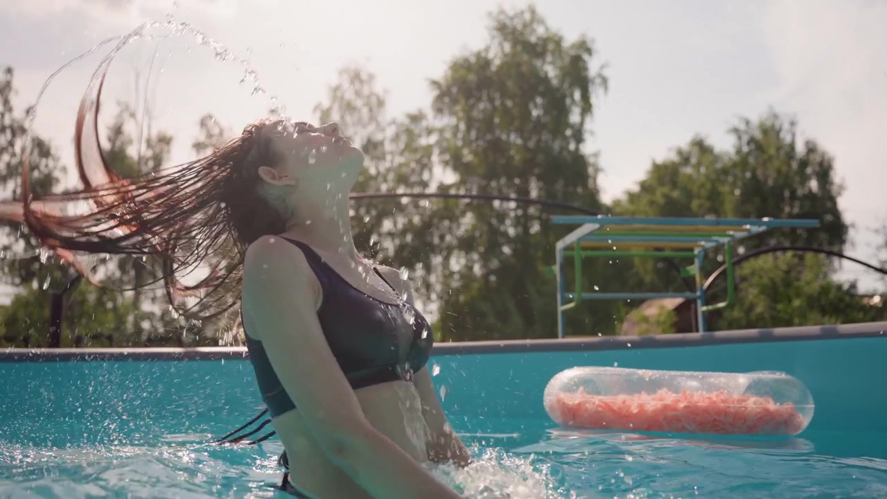 Woman in black swimsuit enjoys water play splashing and relaxing at edge of outdoor pool. scene features bright sunlight clear blue sky green trees beyond fence floating pink objects on calm