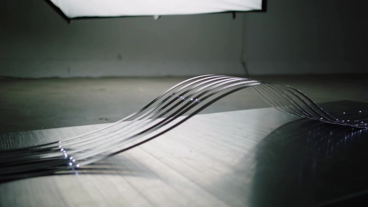 Strisce di metallo si piegano a formare un'onda da sinistra a destra su una scrivania di legno, con un'ancora a sinistra che sposta un softbox proiettando luce.