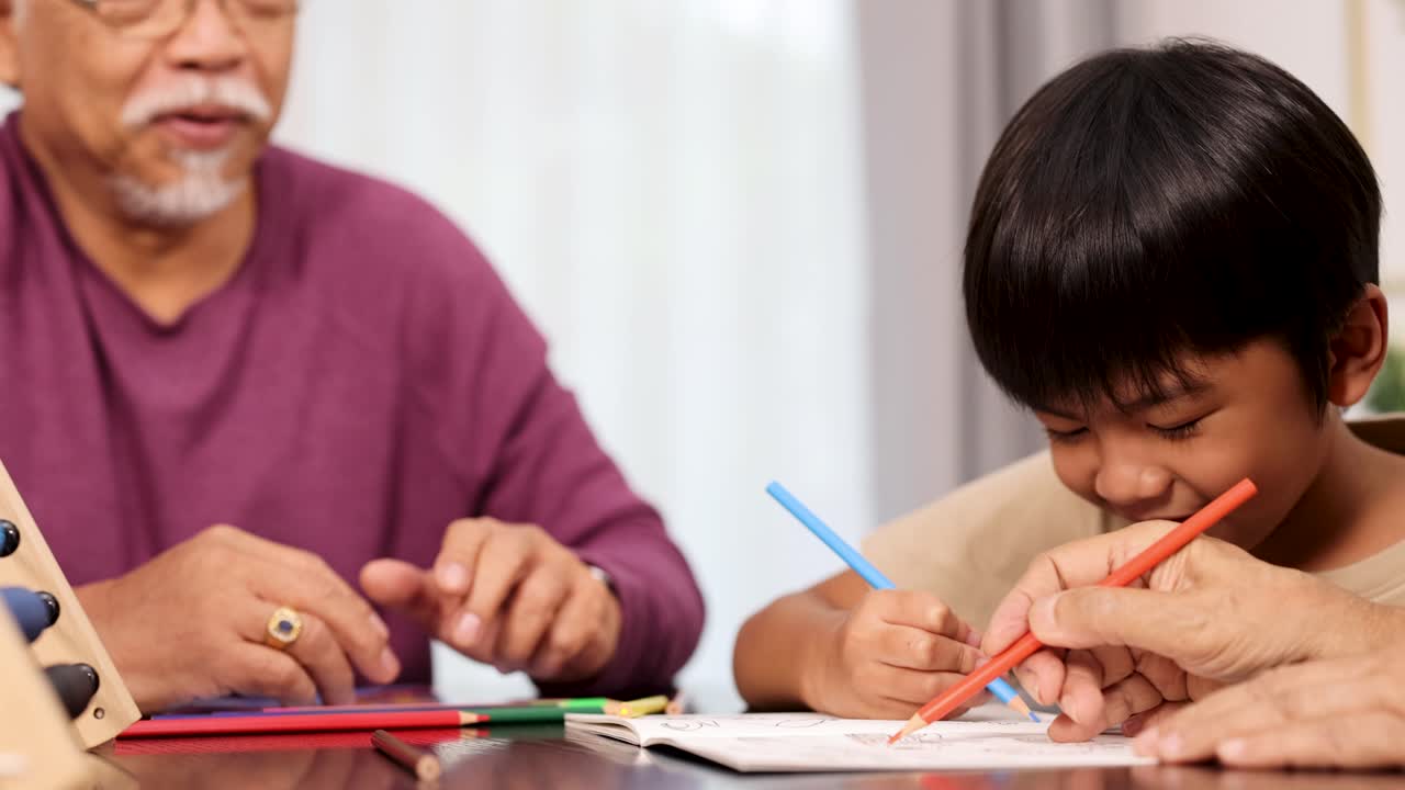 An elderly man and young boy sit at a table, smiling and coloring in a book with colored pencils in a warmly lit living room
