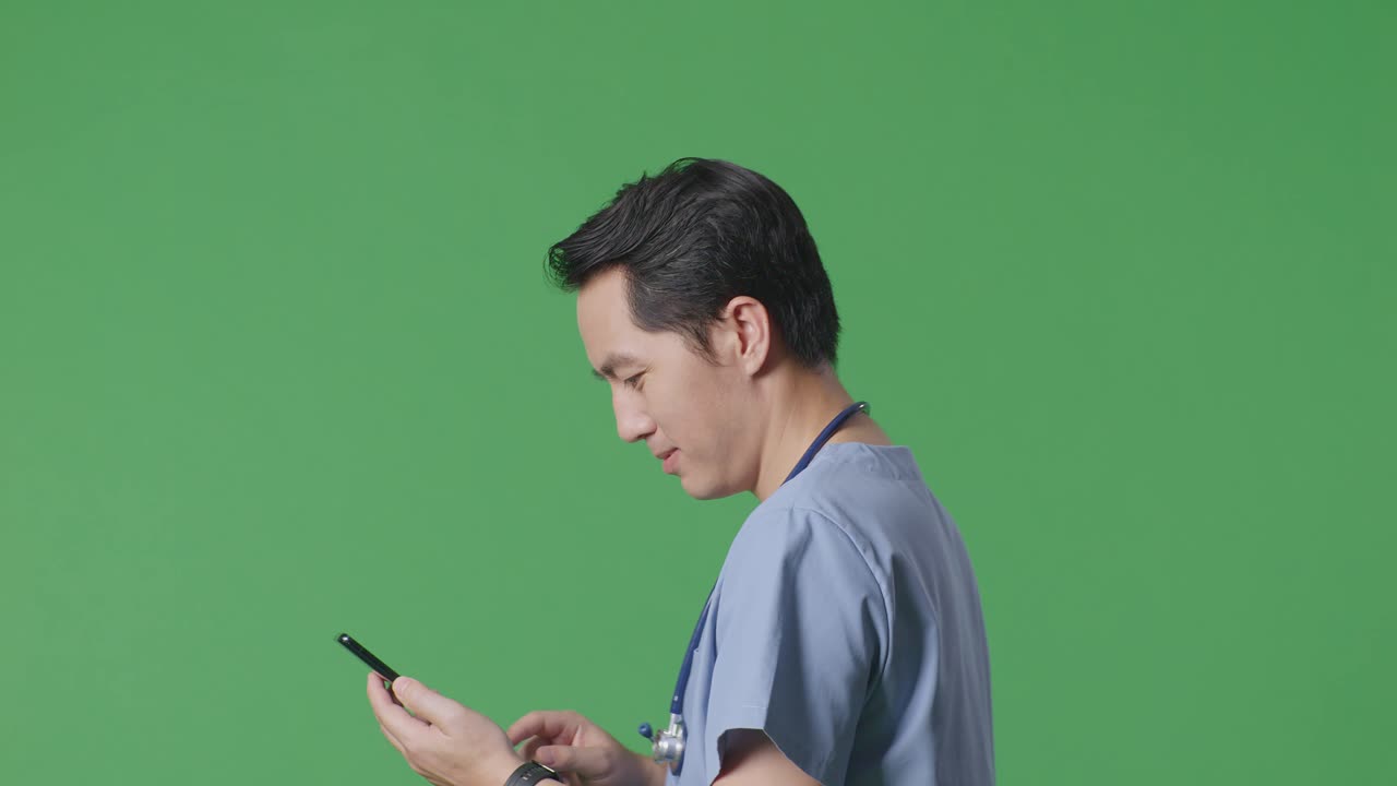 병원 에서 녹색 스크린 배경 에 걸어 다니는 동안 스마트폰 을 사용 하는 스테토스코프 를 들고 있는 아시아 남성 의사