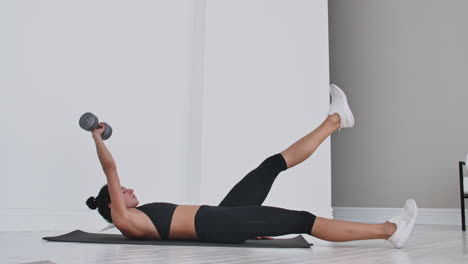 beautiful brunette girl lifts her torso with a dumbbell in her hands to her leg lying on a rug in a white apartment.