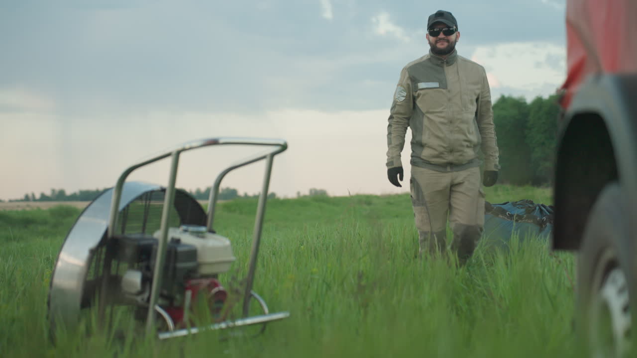 close up of man dragging gear bag across tall grass toward paramotor engine cage at dusk under soft cloudy sky, focus on dynamic posture and movement