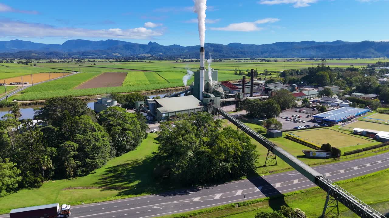 Aerial footage captures a sugarcane factory emitting smoke, surrounded by lush fields and roads under clear blue skies