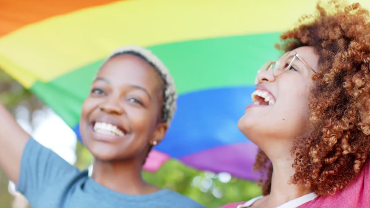 Happy diverse female lesbian couple walking with rainbow flag of pride in slow motion