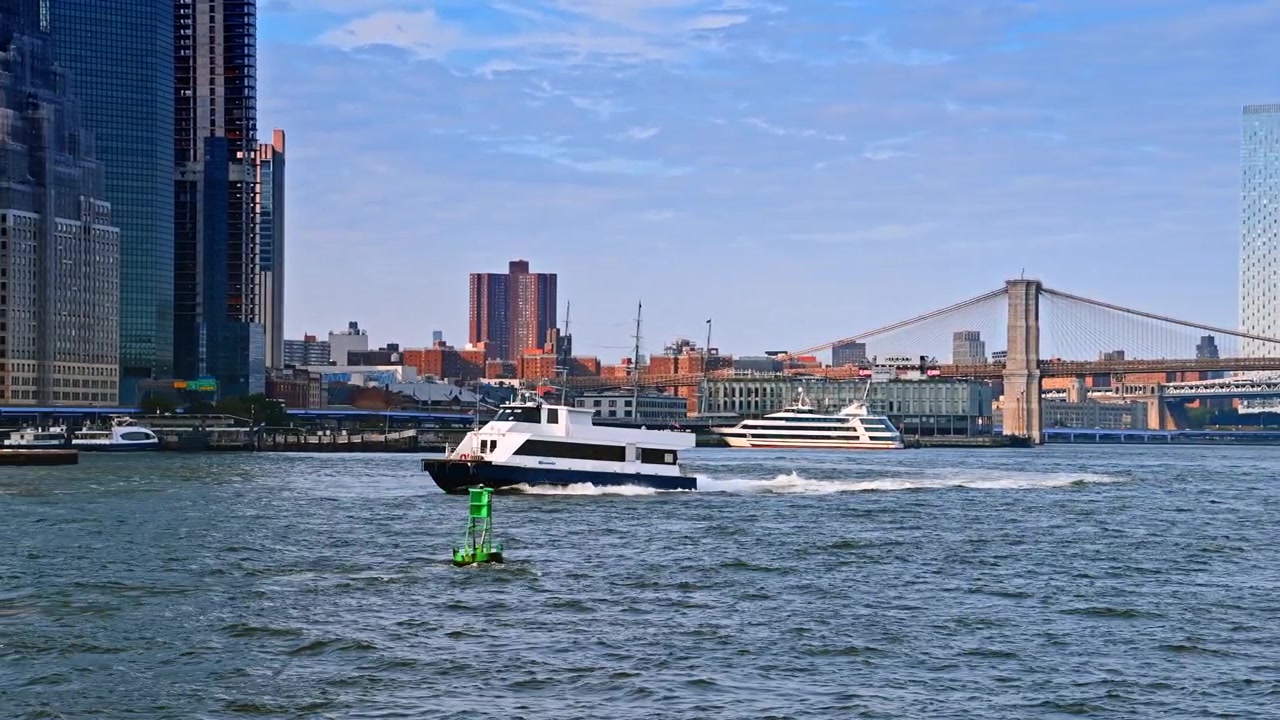 New York, USA, 8 maggio 2026: Traghetto attraversa l'East River vicino al Ponte di Brooklyn, New York City. Un catamarano bianco supera una boa verde con torri e il ponte sullo sfondo.