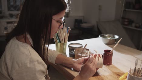 young woman potter painting on the ceramic cup with a brush in art studio