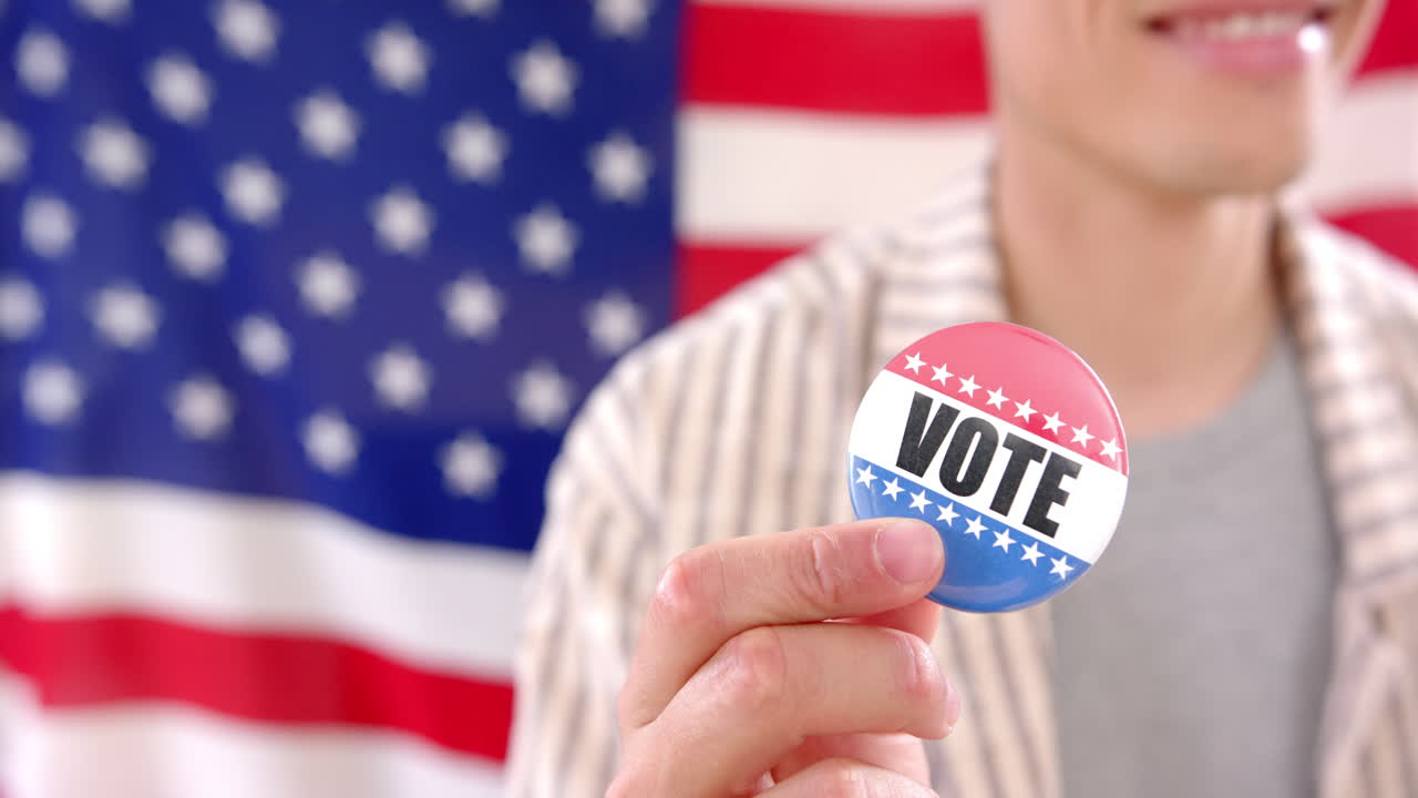 Happy biracial man in front of american flag holding badge with vote text, copy space, slow motion