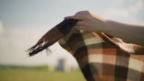 un primer plano de la mano de una mujer, su cara no es visible, sosteniendo una bufanda a cuadros como el viento sopla suavemente en un campo de hierba