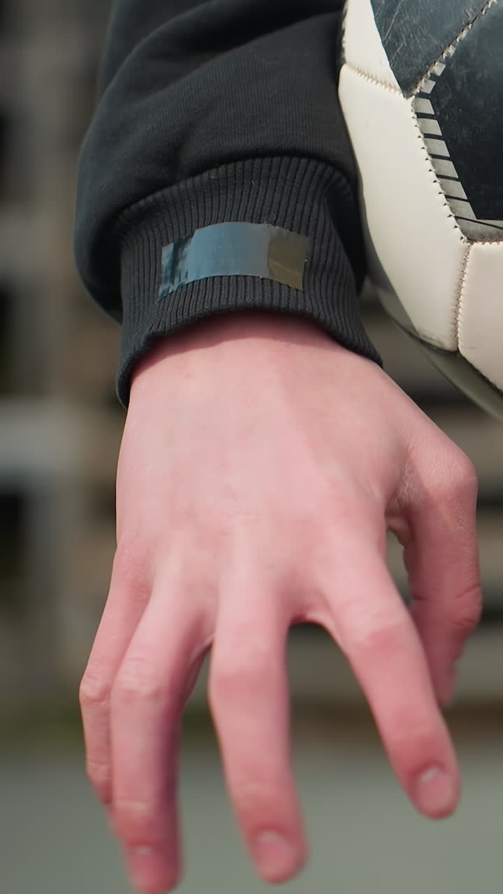 Close-up of an individual holding a worn soccer ball under his arm, as he clasped hand, with a blurred view of a staircase behind