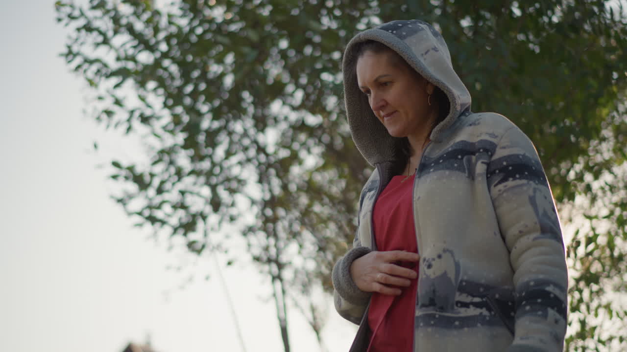Donna bianca incinta che cammina su un sentiero del giardino, cardigan con cappuccio, vestito rosso, mano appoggiata sulla pancia, serra di vetro e casa sullo sfondo, sole mattutino, bagliore caldo, futura mamma giardiniera domestica che controlla le piante.