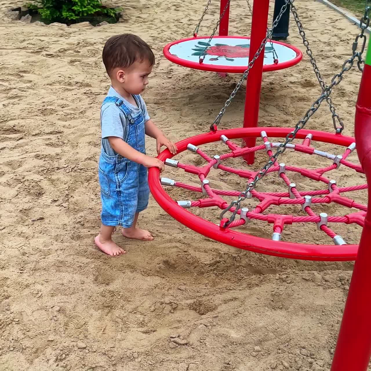 Bebé descalzo con un peto vaquero caminando por la arena en el patio de juegos. Un niño pequeño mueve el columpio de un lado a otro.