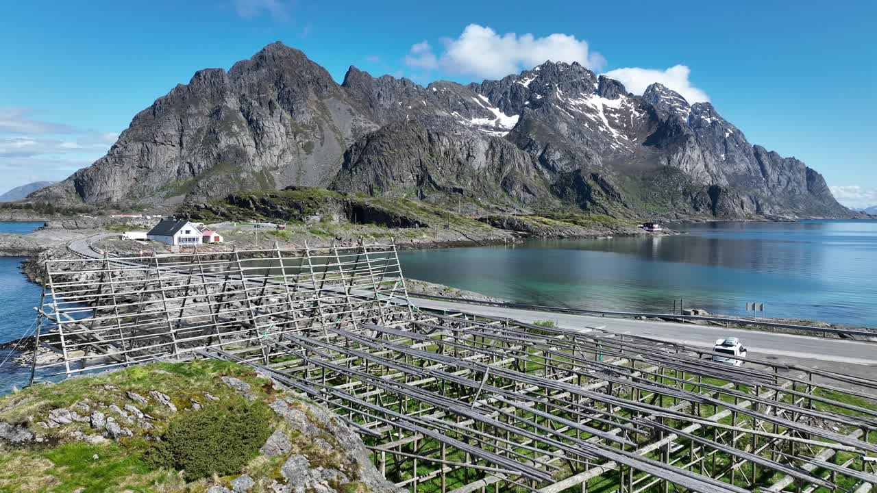 Aerial moves over empty stockfish racks in Henningsvaer with Festvagtinden and peaks beyond