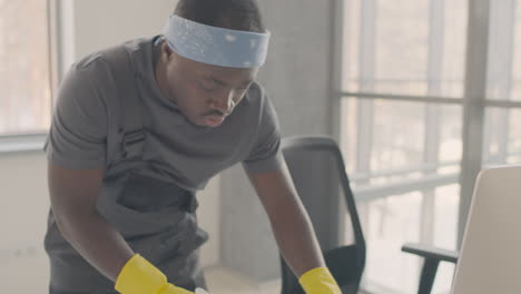 Cleaning-Man-Wearing-Bandana-And-Uniform-Cleaning-A-Desk-Inside-An-Office-1