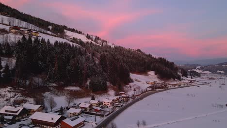 Aerial-shot-of-a-snow-covered-mountain-town,-winding-road-through-the-valley-during-sunset