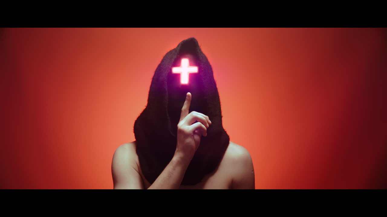 Masked man with cross of light glowing on his face makes a silence gesture - red background