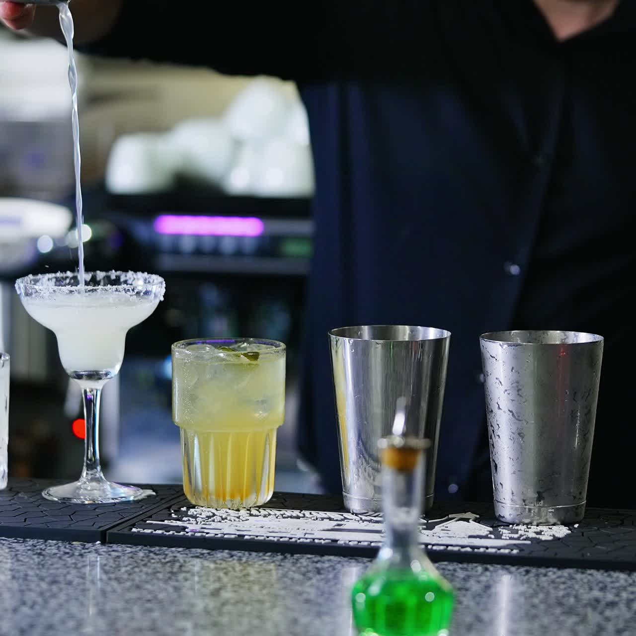 Barman in black shirt filling the wineglass with beverage. Glasses for cocktails making at the bar stand. Close up. Blurred backdrop