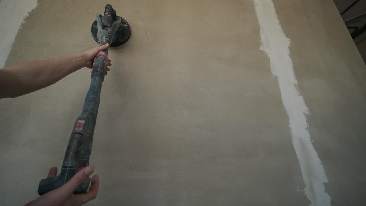 Action shot of construction work, showing a hand guiding a power sander on a wall, part of the process of smoothing surfaces for a fine finish in renovation