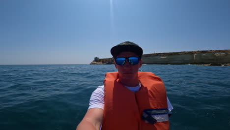 young man in self-portrait with life jacket on a boat tour in malta with ocean and cliffs in the background