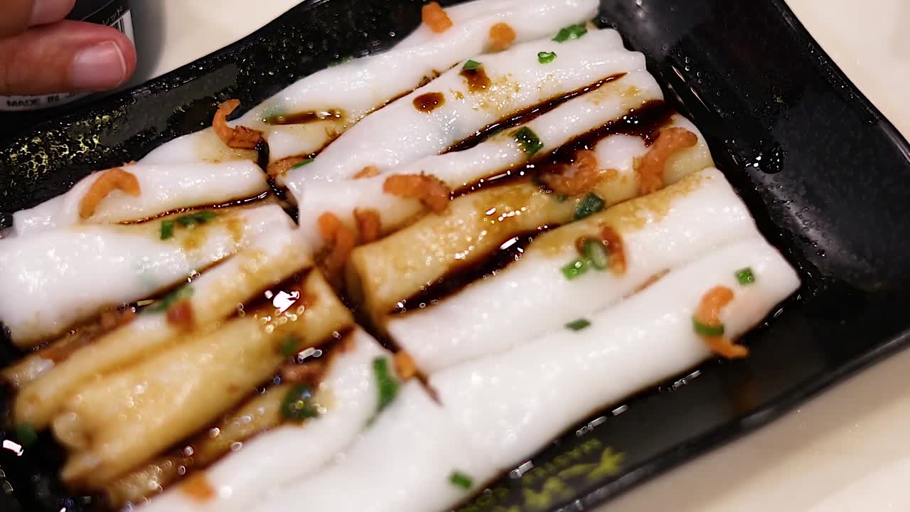Close-up of sauce being poured over rice noodle rolls on a black plate.