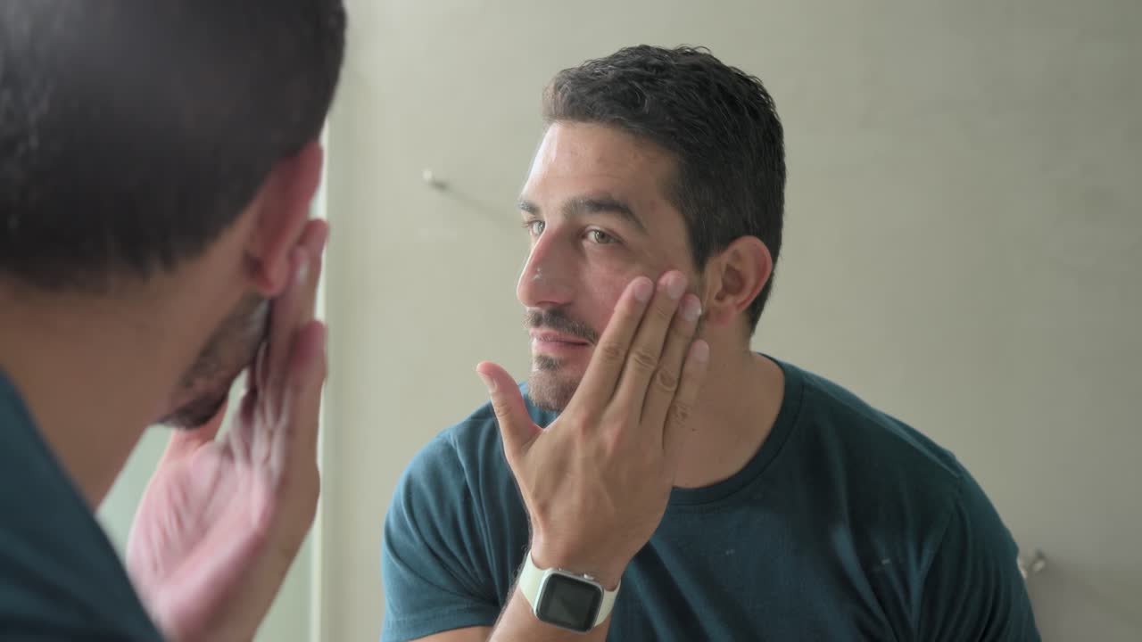 Uomo adulto si china allo specchio controllando la pelle per la cura, tamponando e strofinando la crema sul mobiletto del bagno.