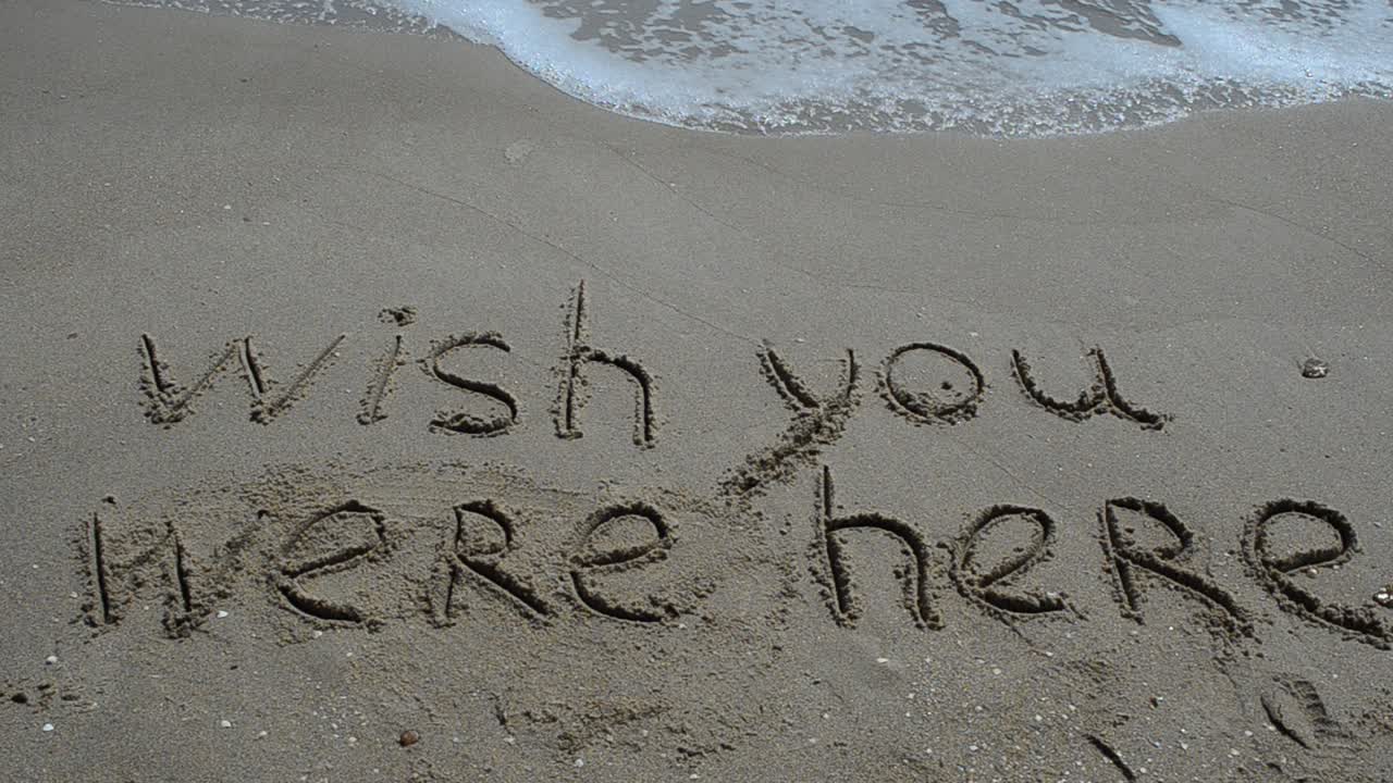 Testo concettuale 'wish you were here' scritto a mano sulla sabbia di una spiaggia.