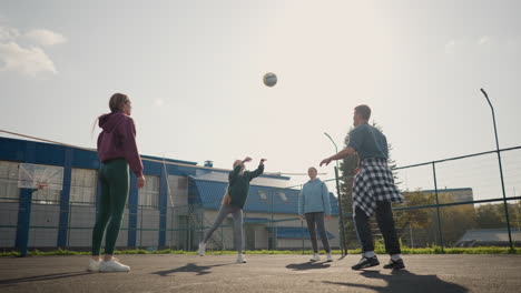 el entrenador guía la sesión de entrenamiento con tres damas, pasando la pelota de voleibol entre ellas en la cancha al aire libre, la escena muestra el trabajo en equipo y la práctica con el edificio y el fondo de la valla