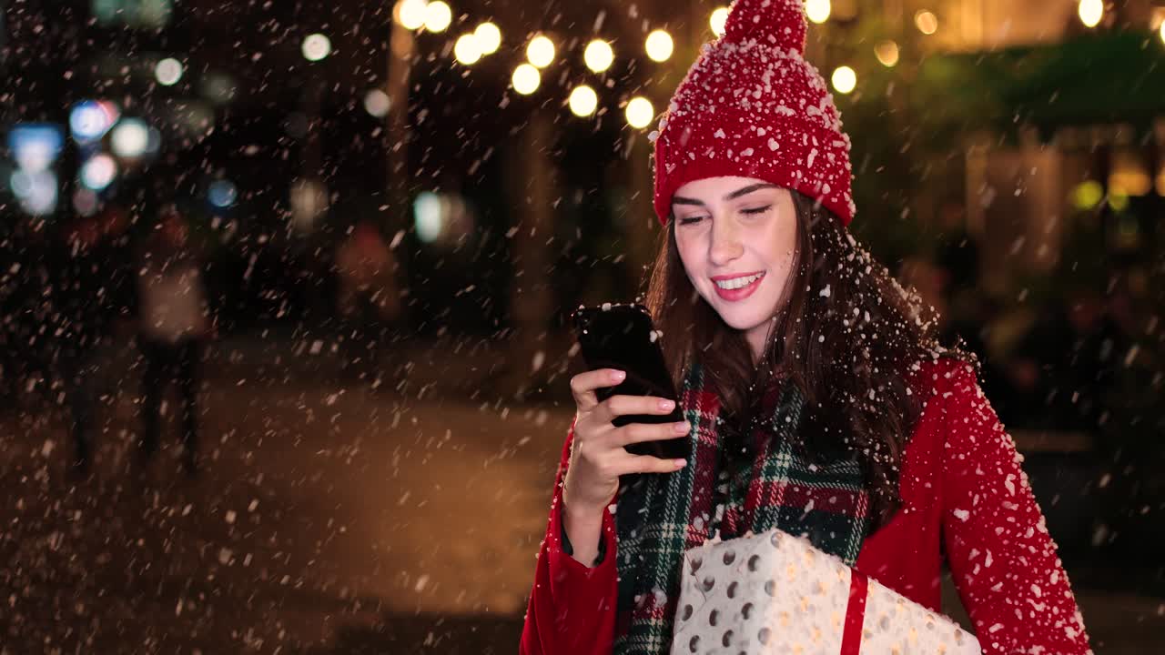크리스마스에 눈이 내리는 동안 거리에서 선물을 들고 스마트폰을 사용하는 빨간 코트를 입은 백인 여성의 클로즈업 보기