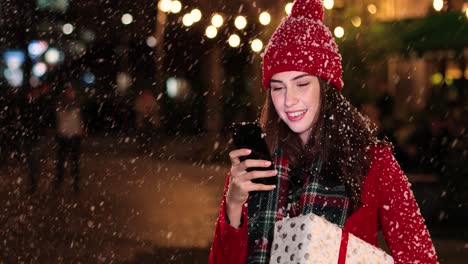 close-up view of caucasian woman in red coat holding a present and using smartphone on the street while it‚äôs snowing in christmas