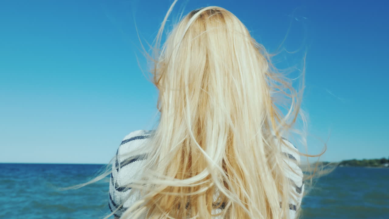 긴 머리를 가진 금발의 여자는 바다 바람을 바라보며 머리를 헝클어뜨린다