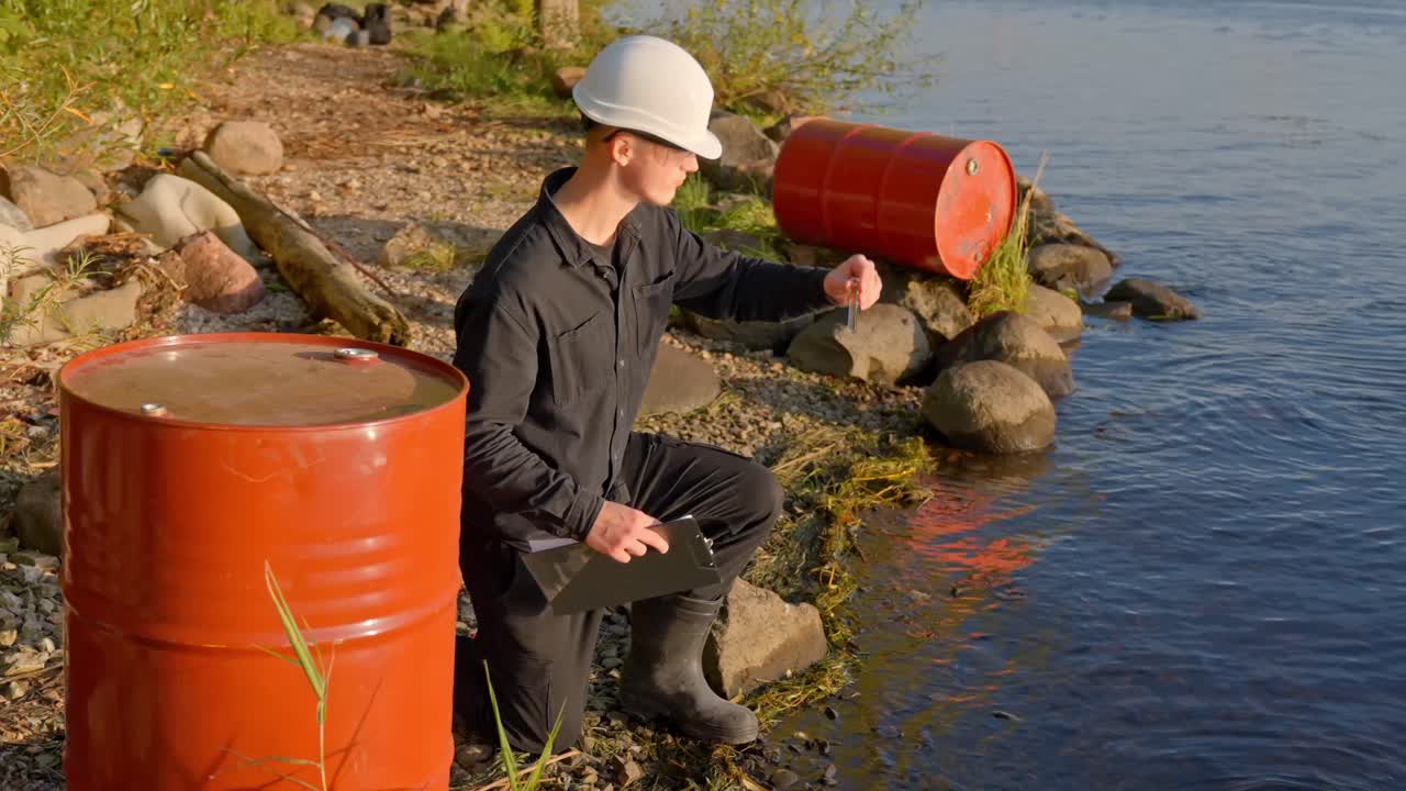 흰색 안전모와 유니폼을 착용한 현장 과학자가 강둑 근처의 주황색 산업용 통 옆에서 물 샘플을 수집하고 관찰하고 있어요. 점검 결과 환경 오염 위험이 강조되었어요.