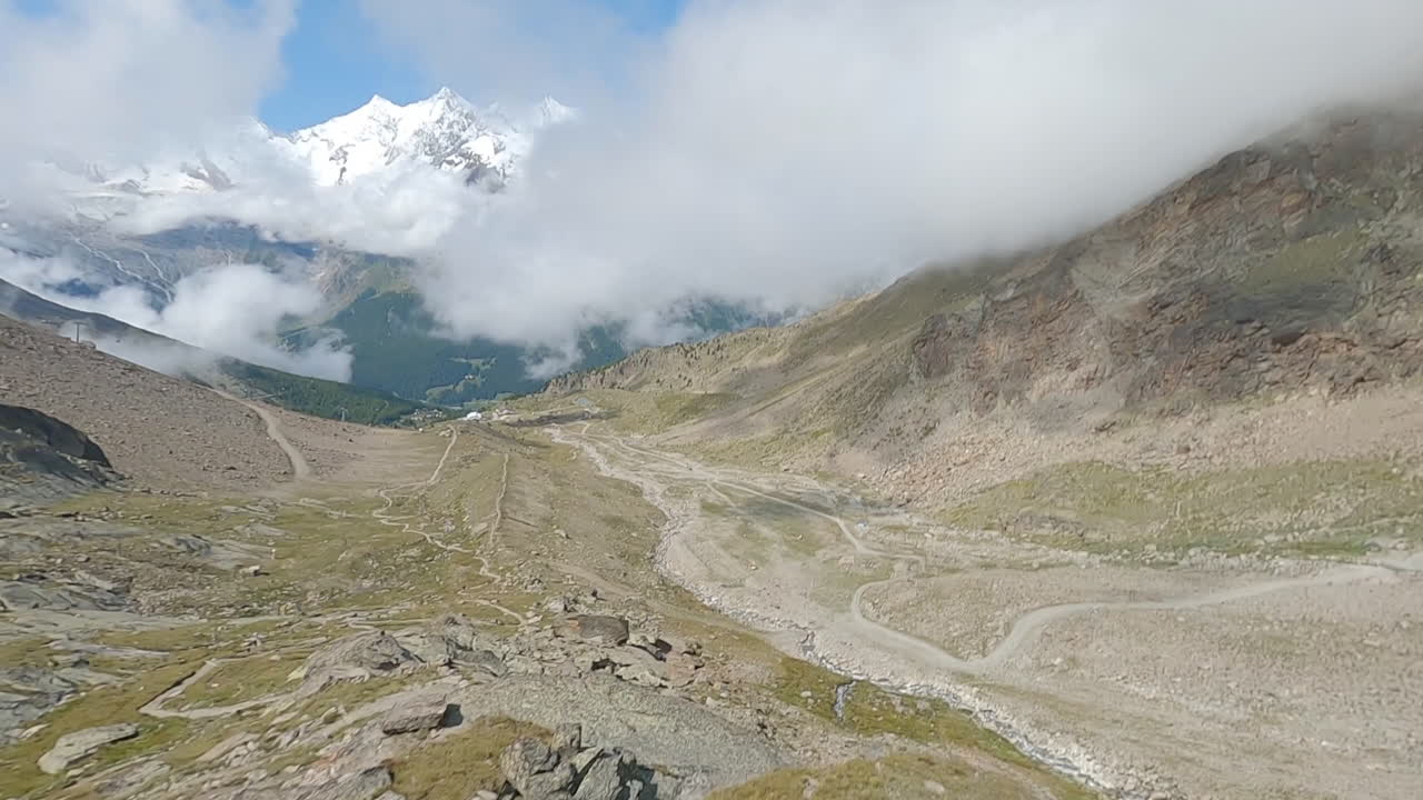 FPV drone follows two mountain bikers descending steep, exposed alpine trails above Saas-Grund and Saas-Fee on a clear bluebird summer day, showcasing epic terrain and speed