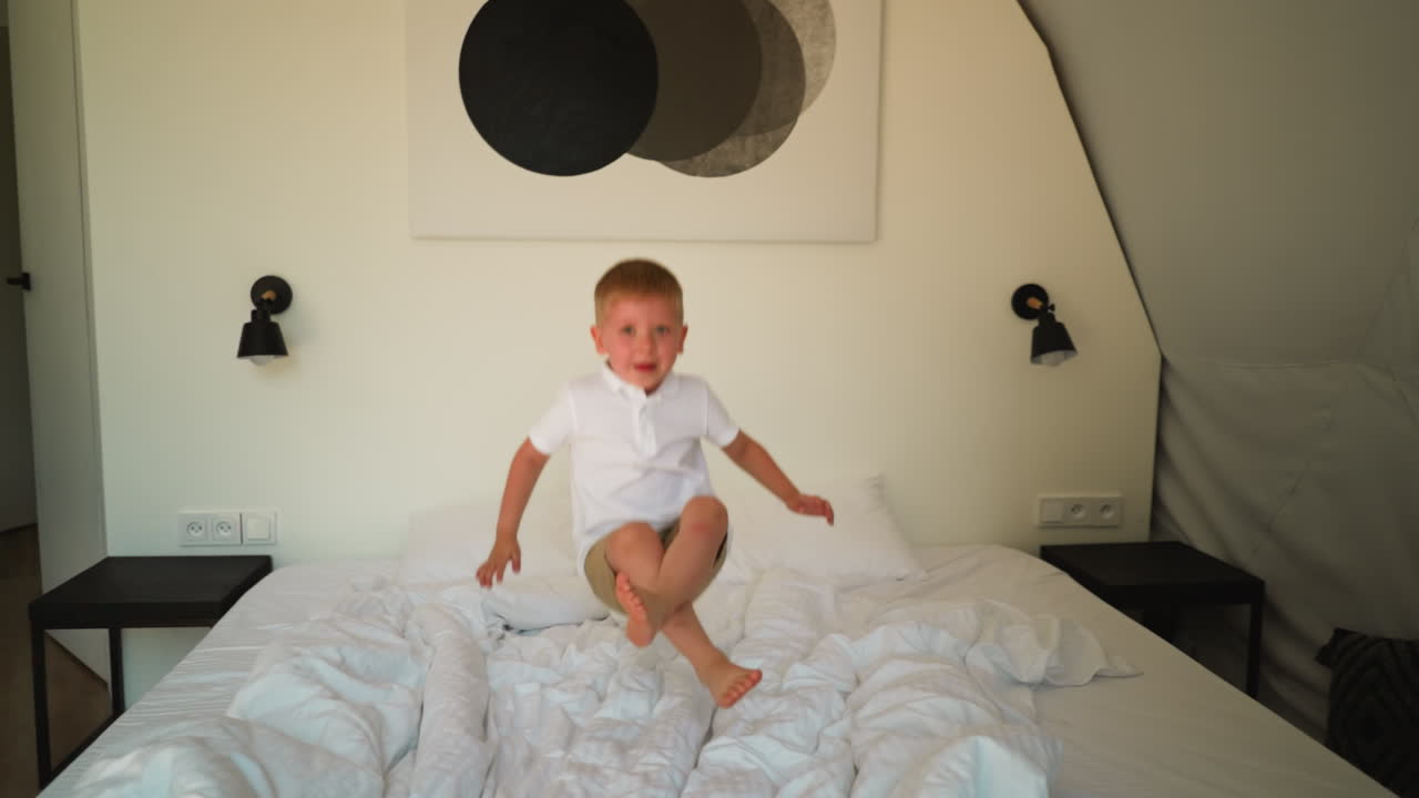귀여운 소년은 밝은 색의 린으로 침대에 뛰어들어, 유쾌한 유아는 현대적인 가운 침실에서 부드러운 침대에 뛰어다니며 좋은 시간을 보내고, 행복한 어린 시절의 순간.