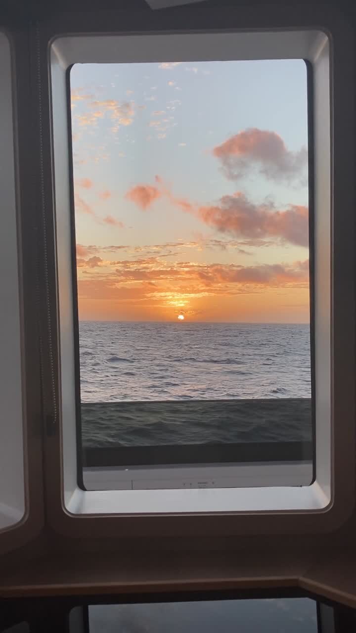 Filmato verticale da telefono del sole che tramonta all'orizzonte, visto attraverso i finestrini del ponte di un grande yacht in navigazione.