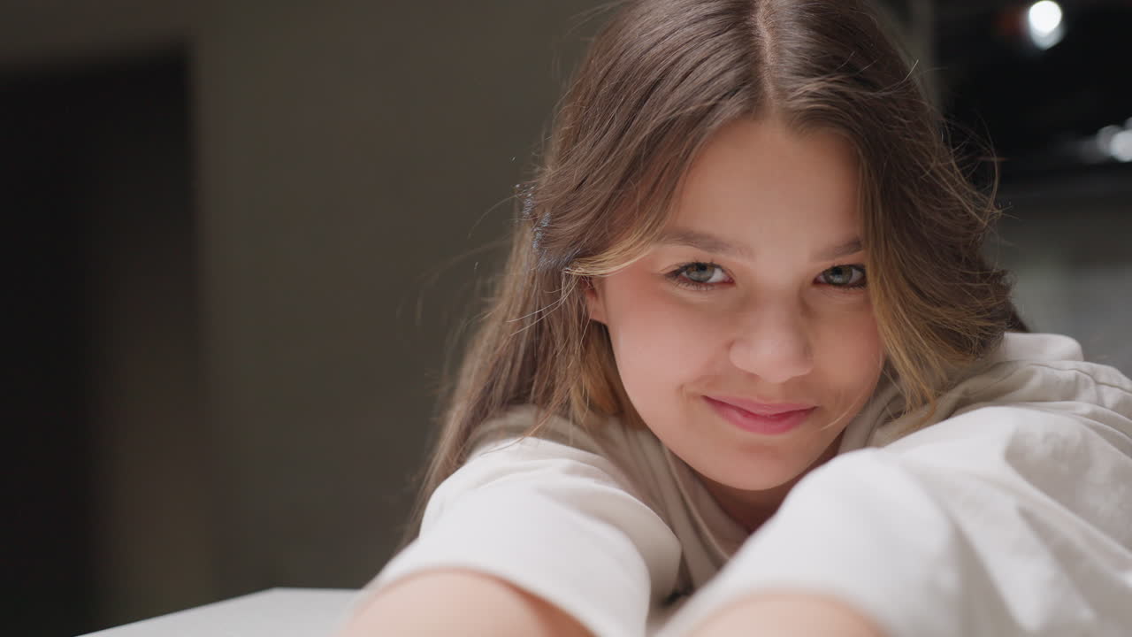 Portrait of cheerful young lady seated at table stretching forward with hands resting on surface, casual indoor moment showing relaxation, natural beauty in warm lifestyle environment with soft light