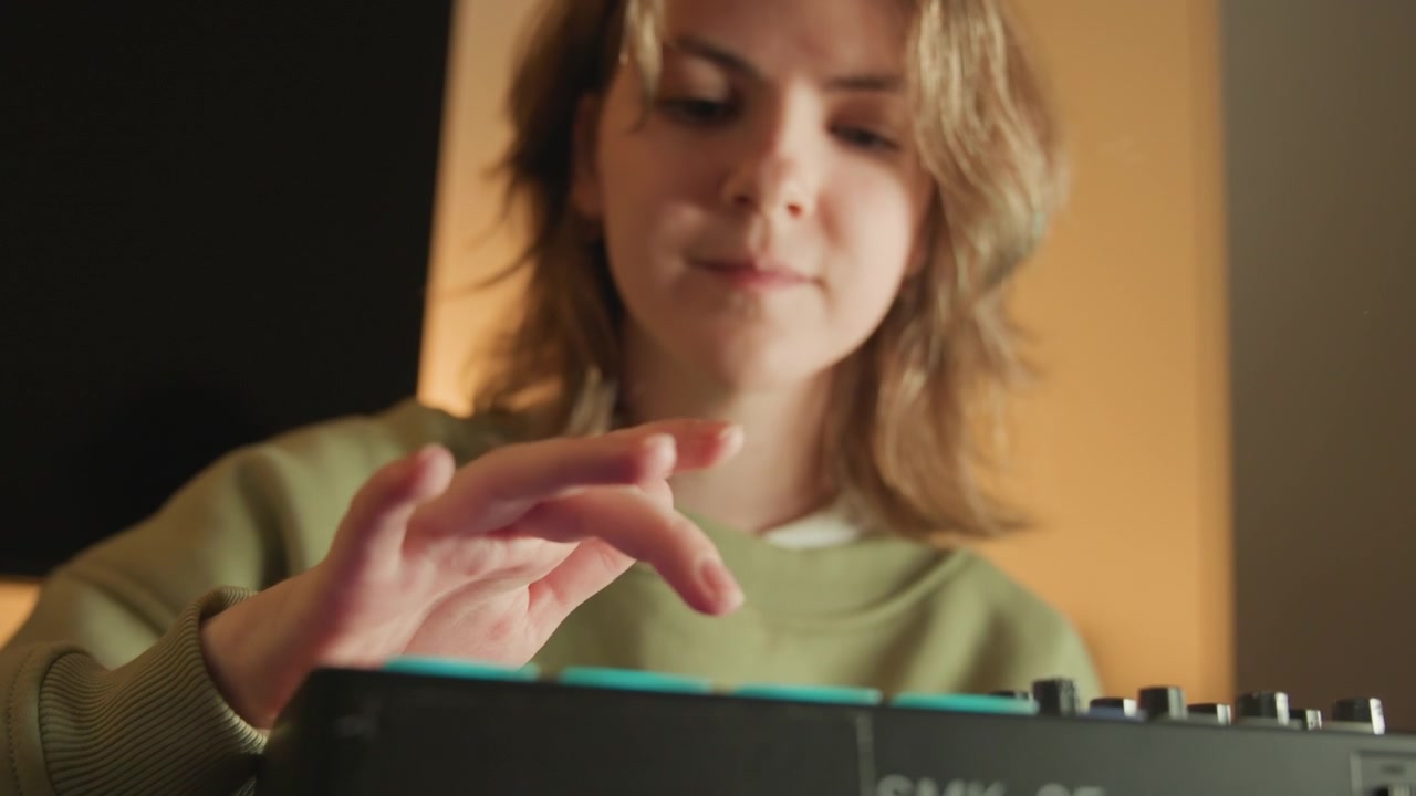 Joven con sudadera verde presiona teclas en un teclado electrónico mientras sus dedos se mueven con un desenfoque visible, creando una escena interior tranquila de práctica concentrada, ejecución suave y concentración natural