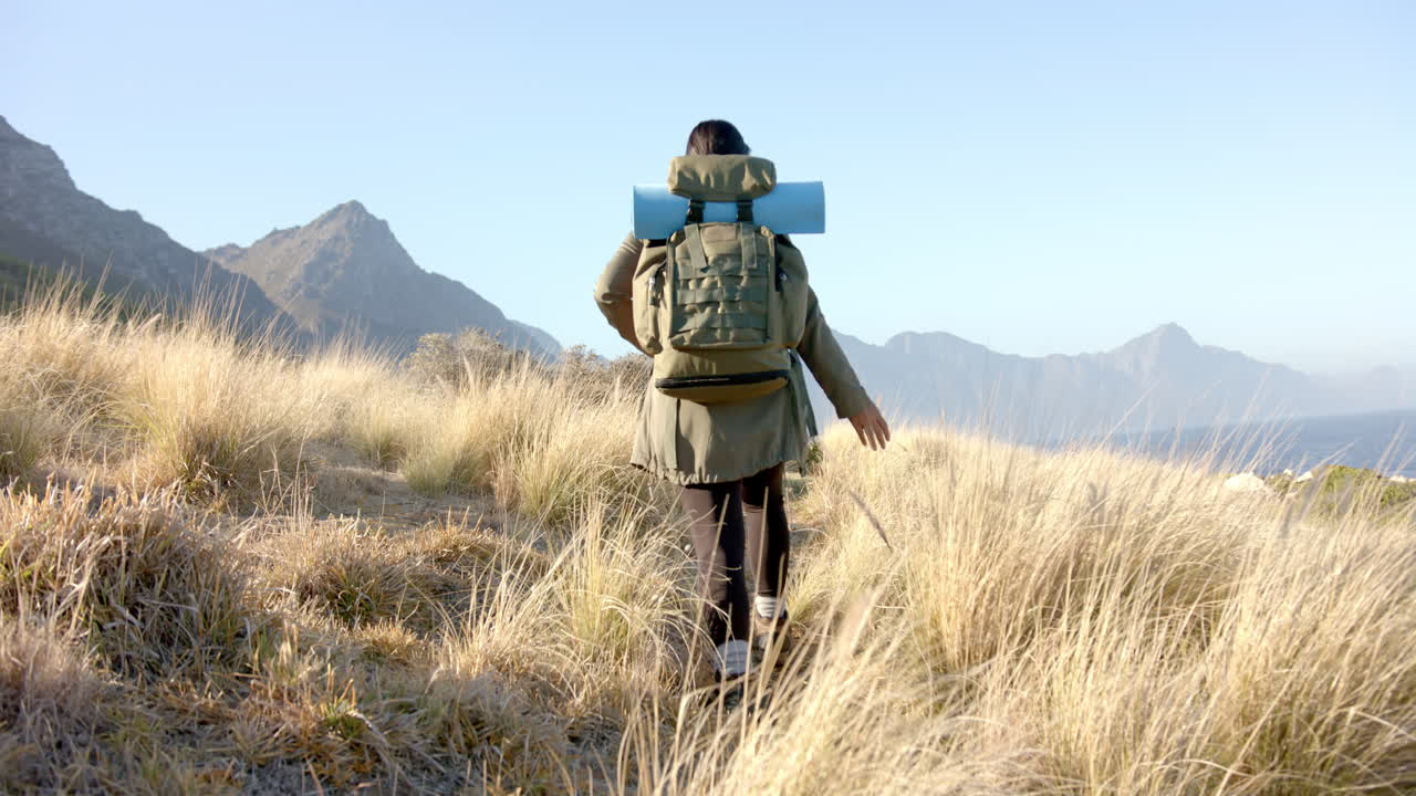 Hiking in mountains, woman with backpack and camping gear walking through grassland