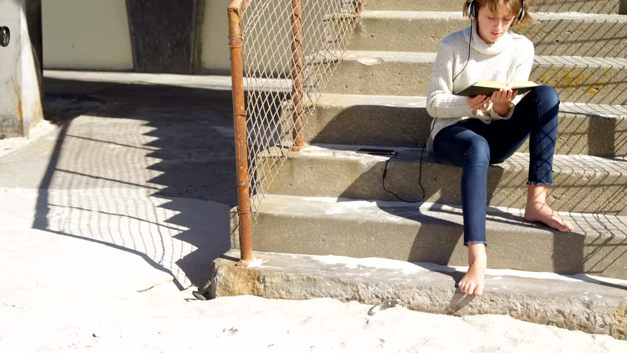 해변 4k에서 책을 읽는 동안 음악을 듣는 소녀