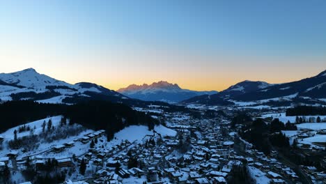 the drone is flying over a village in a valley covered in snow with big mountains in the background and the sun setting in austria aerial footage 4k