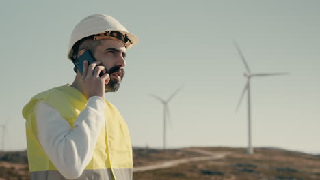 Nahaufnahme-Eines-Kaukasischen-Ingenieurs-In-Einem-Feld-Mit-Windkraftanlagen,-Der-An-Einem-Sonnigen-Tag-Einen-Anruf-Entgegennimmt