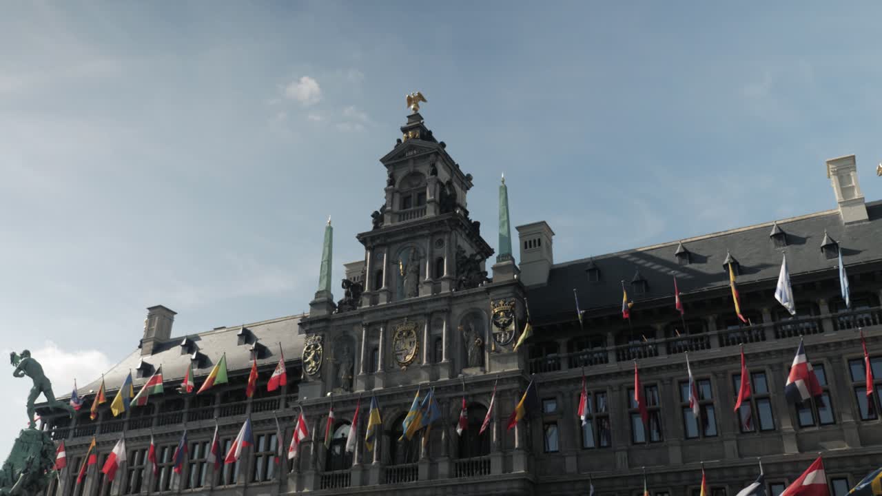 Slow motion push-in shot of Antwerp City Hall in Belgium, showcasing its ornate Renaissance architecture adorned with international flags, under a bright blue sky