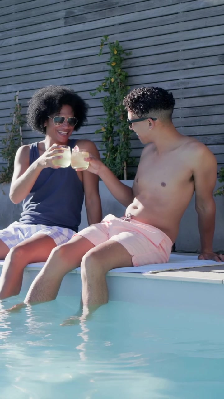 Vertical video: Smiling friends clinking drinks, sipping and talking poolside in trunks and shades