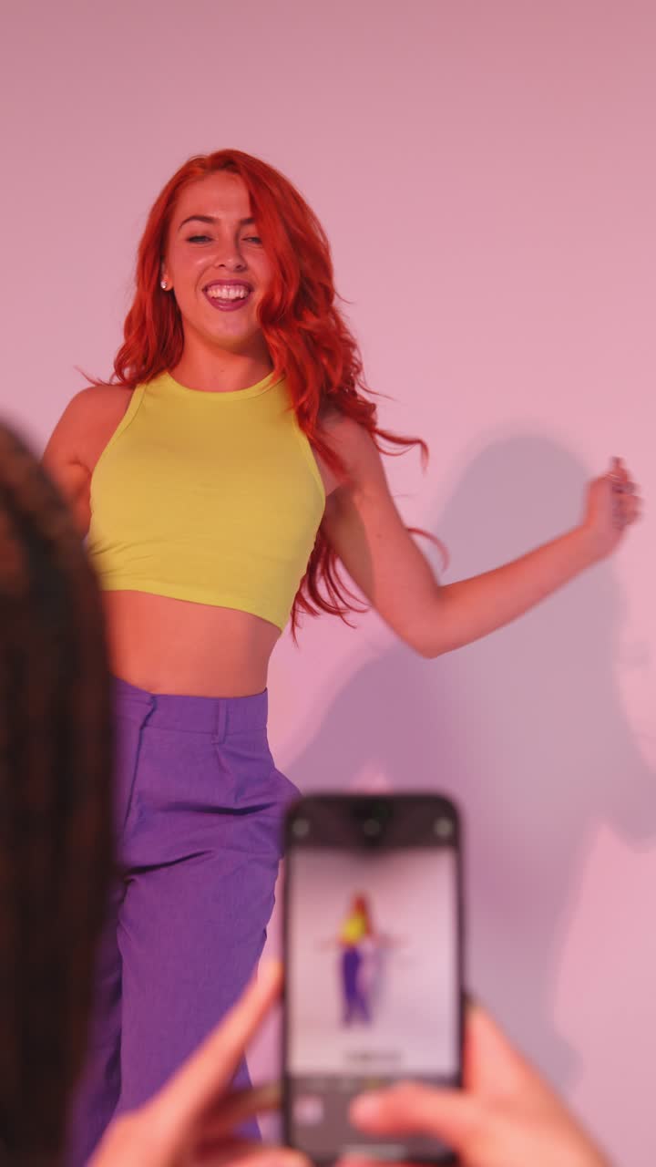 video studio verticale di una donna che scatta una foto di un'amica che balla sul cellulare contro uno sfondo rosa 1