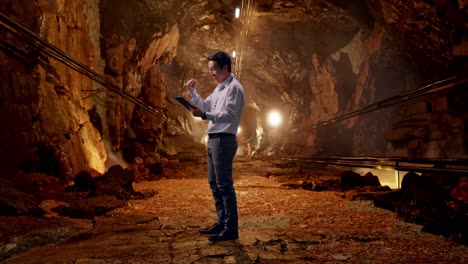 Full Body Side View Of An Asian Male Professional Worker Standing With His Tablet In Underground Mine Tunnel, He Raises His Fist Up With Screaming Goal