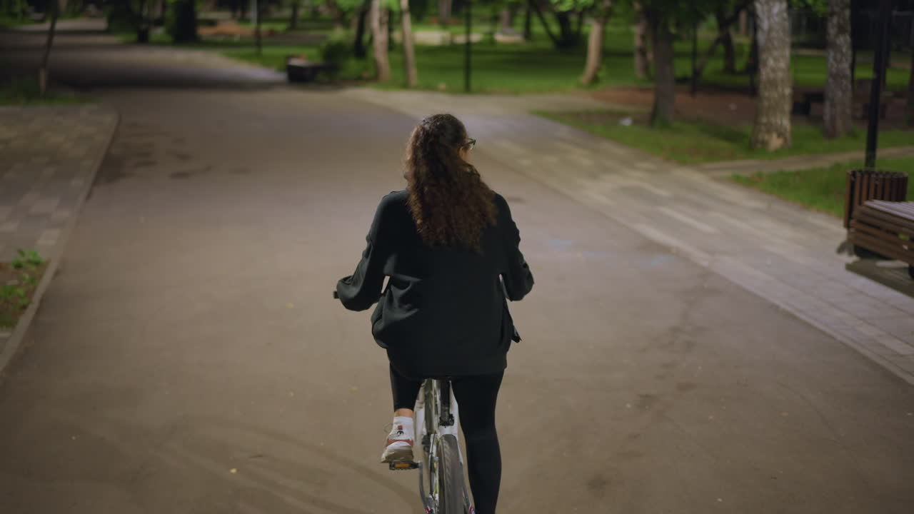 Woman Pedals Through Tranquil Park, Serene Young Lady Rides At Night Quietly, Calm Female Cyclist On Moonlit Park Trail Alone, Peaceful Young Woman Journeys Through Dimly Lit City Park At Night