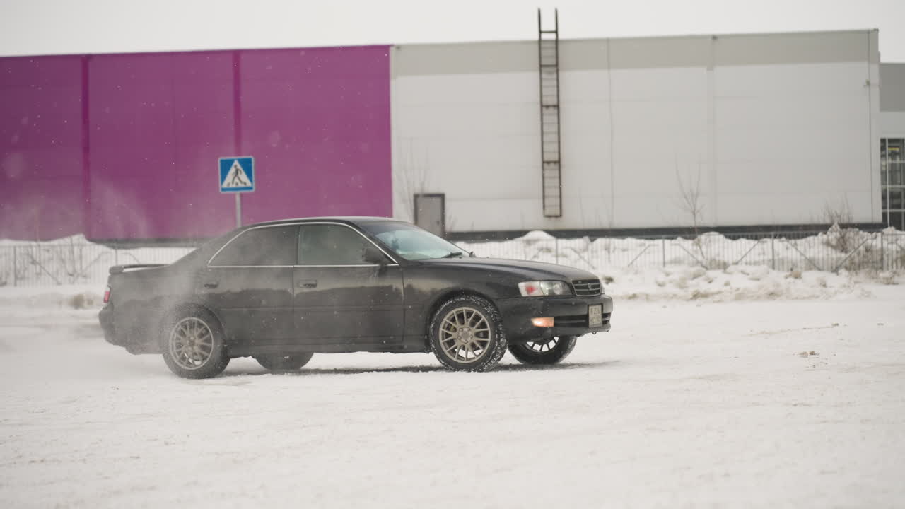 car drifting and rotating on snowy ground during winter with snow mist swirling around tires in front of modern industrial building and purple wall background