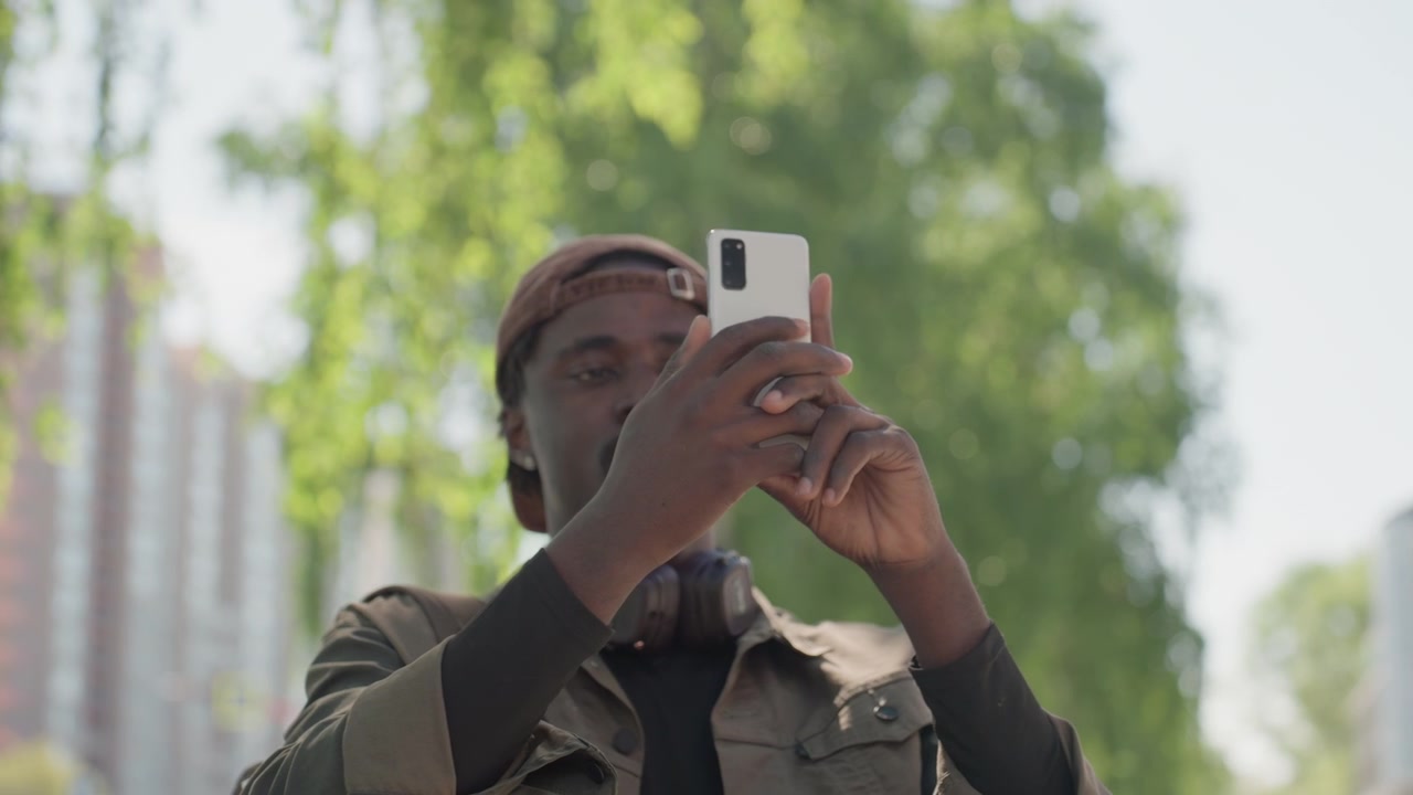 Gros plan d'un homme tenant un smartphone pour un selfie dans un cadre urbain extérieur avec des arbres verts et des bâtiments visibles. L'homme porte une casquette marron et une veste par-dessus une chemise noire, l'angle de la caméra reste stable.