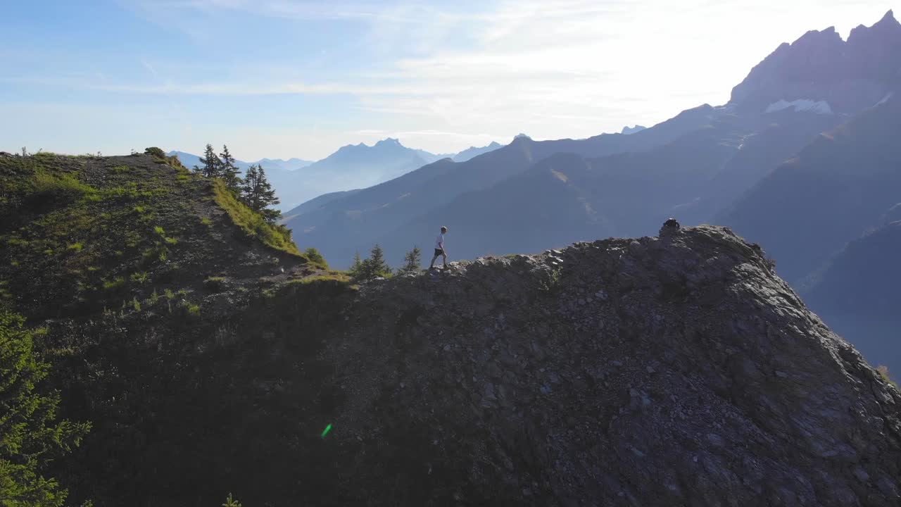 Drone shot of hiker standing on rocky mountain ridge in Canada surrounded by vast landscape, green forest, and distant peaks under clear sky in peaceful natural environment during daylight