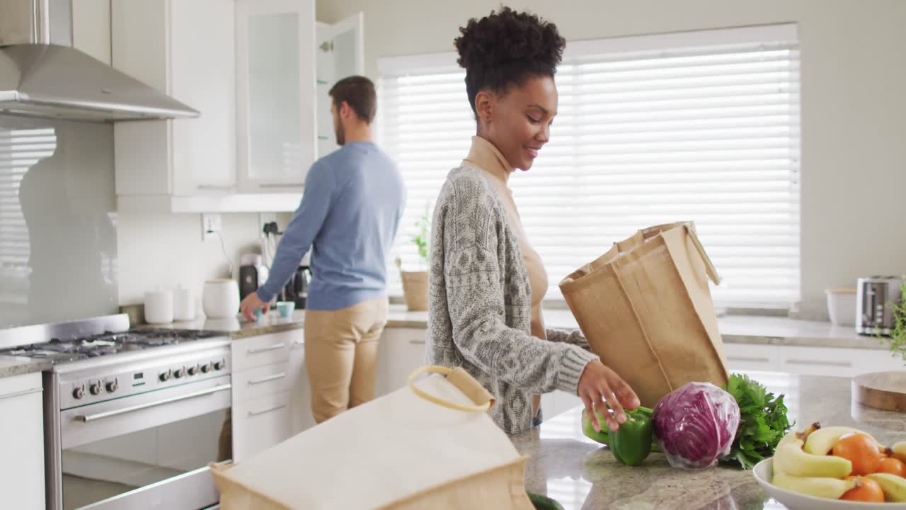 Video of happy diverse couple unpacking groceries in kitchen