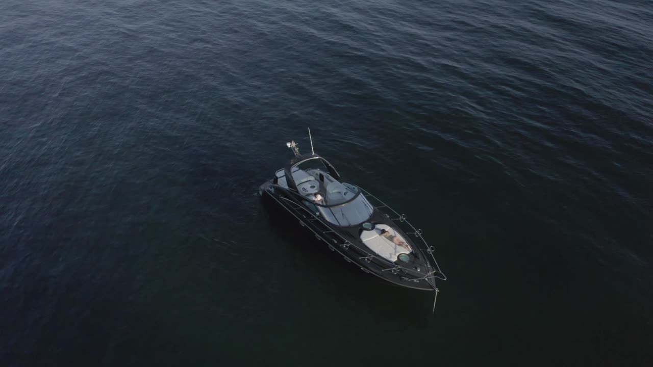 Yacht Floating And Anchored On The Calm Sea. Man Stands Behind The Steering Wheel With A Girl In Swimsuit Lying And Sunbathing On The Deck Of A Yacht In Albufeira, Portugal. - aerial reveal shot
