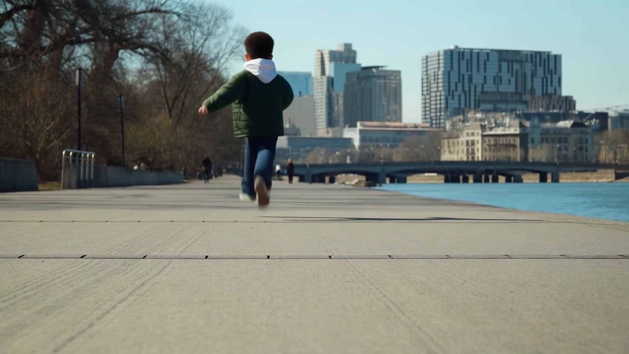 Low rear view shows a child running steadily toward a bridge beside blue water, with long concrete walkway, bare trees, scattered pedestrians, and modern buildings under clear daylight.
